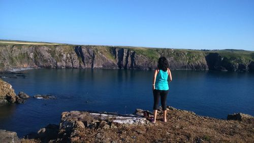 Rear view of woman standing on rock by sea against clear blue sky