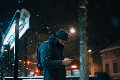 Man using mobile phone while standing against building during snowfall at night