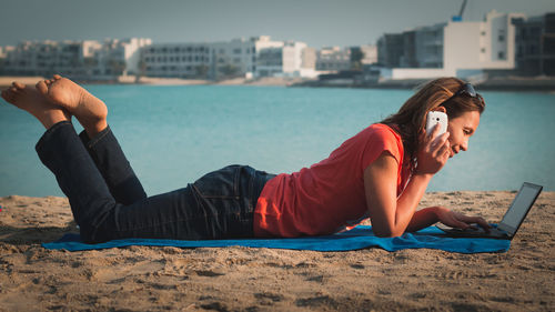 Woman talking on mobile phone while working on laptop at beach