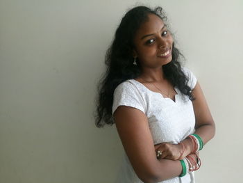 Portrait of smiling young woman wearing tricolor bangles standing with arms crossed against wall