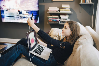 Side view of girl taking selfie through mobile phone while using laptop on sofa