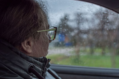 A girl with glasses sits in a car and looks out the side window. there are raindrops on the glass