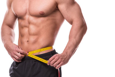 Midsection of shirtless man measuring abdomen against white background