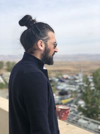 Side view of young man holding coffee cup while standing against sky