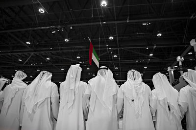 Rear view of men wearing white traditional clothing with united arab emirates flag hanging from ceiling in background