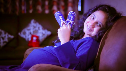 Portrait of pregnant woman holding shoes while resting on sofa