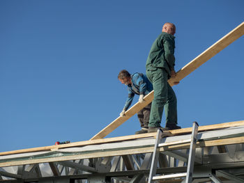 Colleagues adjusting wooden plank on roof against clear sky