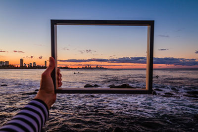 Cropped hand holding frame by sea during sunset