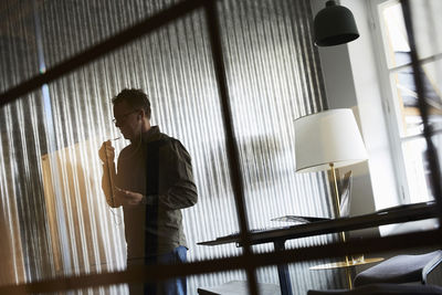 Tilt shot of businessman talking on smart phone through headphones seen from glass at creative office