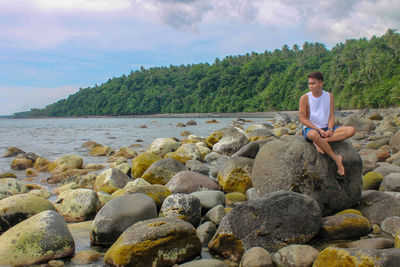 Man sitting on rock while looking at sea against sky