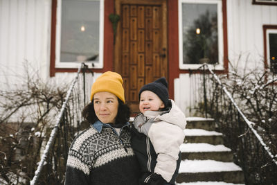 Smiling mother carrying daughter in front of house while looking away during winter