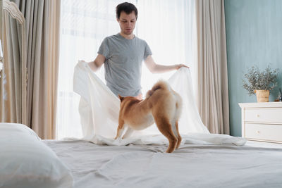 Young man and his funny dog are putting the bedding cover or mattress pad on the bed