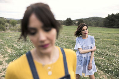 Positive female friends walking in field covered with grass and small delicate flowers near woodland and mountains under clear sky in daytime