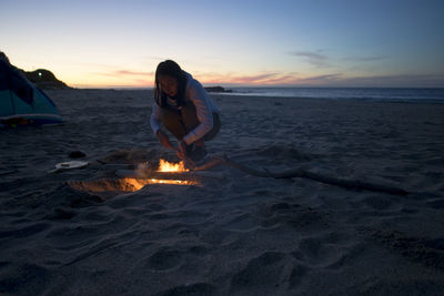 Woman standing by campfire on beach against sky during sunset