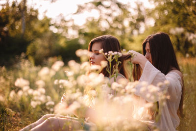 Young woman tying girlfriend’s hair sitting amidst flowers on meadow