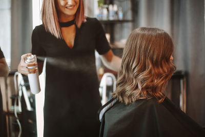 Smiling woman spraying beauty product on hair of customer at salon
