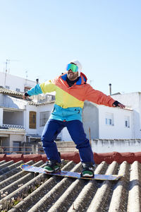 Young guy with cap and goggles smiling with open arms snowboarding on a roof