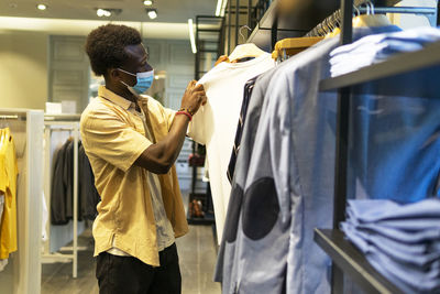 Side view of african american male in medical mask standing in clothing store and choosing t shirt during coronavirus epidemic