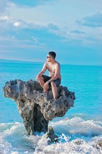 Young man on rock in sea against sky