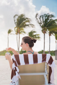 Rear view of woman having drink while sitting on lounge chair at beach against trees