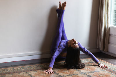 Full body of graceful young hispanic female with long dark hair in sportswear performing asana with raised legs on wall while practicing yoga in cozy apartment