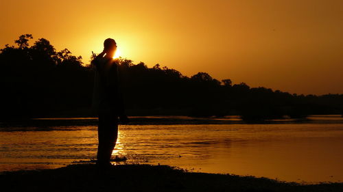 Silhouette person standing by lake against sky during sunset