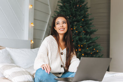 Young asian woman in cozy white knitted sweater using laptop on bed in room with christmas tree