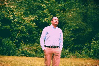 Thoughtful man with hands in pockets standing on grassy field against trees