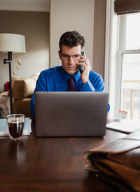 Man on cellphone working from home using a computer at a dining table.