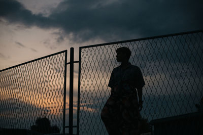 Silhouette woman standing by fence against sky during sunset