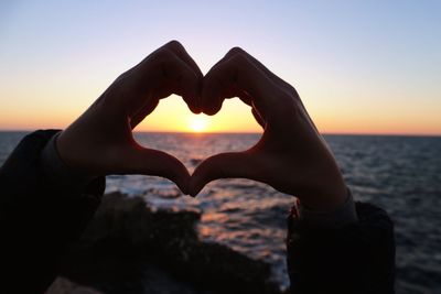Close-up of hand holding heart shape against sky at sunset