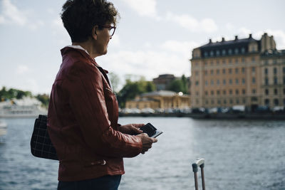 Side view of senior woman holding mobile phone while standing by sea in city