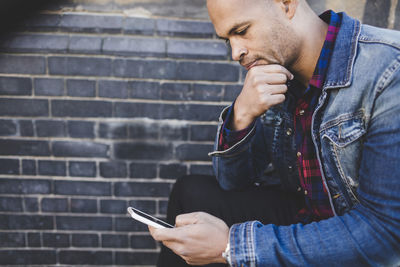 Mid adult man using mobile phone while sitting against brick wall