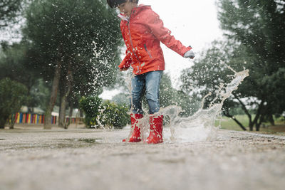 Adorable joyful child in red raincoat and rubber boots having fun jumping in puddle on street in park in gray day