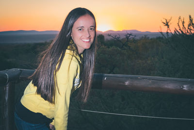 Portrait of smiling young woman standing against railing during sunset