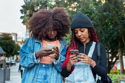 Young ethnic female friends with afro hairstyles concentrating while standing on blurred city street together and browsing mobile phones