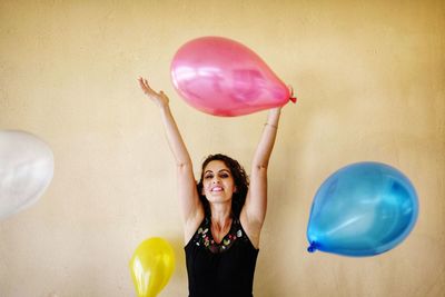 Portrait of smiling young woman playing with balloons while standing against wall