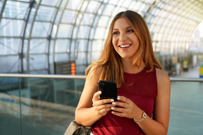 Confident woman looking to the side holding smartphone inside of modern airport. copy space.