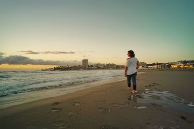 Rear view of man walking at beach against clear sky during sunset