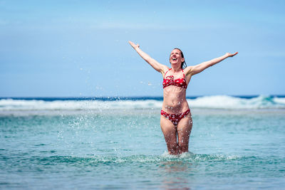 Happy mid adult woman with arms outstretched in sea against sky