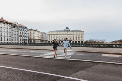 Happy young romantic couple in stylish clothes laughing and holding hands while crossing bridge with historic buildings in background during city tour in bayonne in france