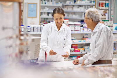 Smiling pharmacist holding prescription in front of man in pharmacy