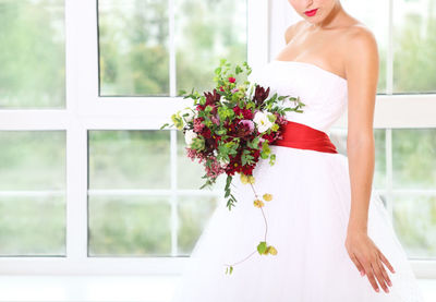 Midsection of bride holding bouquet standing against wall