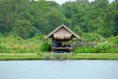 Woman sitting in hut by lake against trees