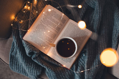 Cup of fresh black tea stay on open paper book with knit cloth in chair over glow christmas lights 