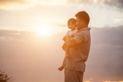 Rear view of father holding hands while standing against sky during sunset