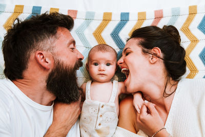 From above serene baby lying on bed among mother and father cuddling and pretending screaming while teasing child