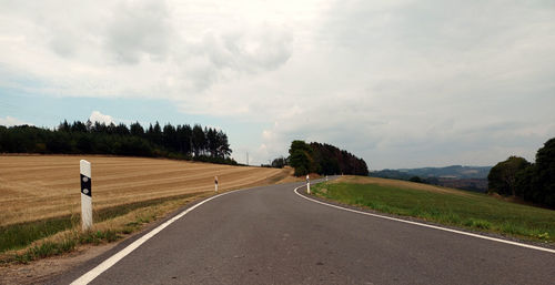 Empty small road against sky through nature with fields and trees in the background.