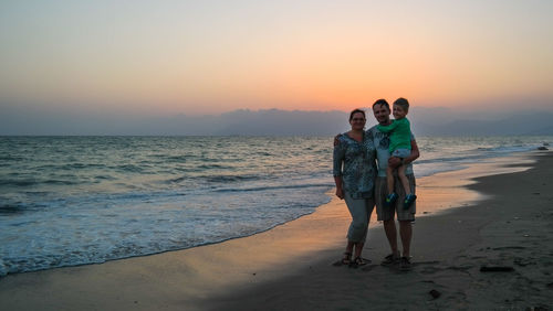 Family standing at beach against sky during sunset