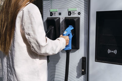 Young woman holding electric plug at charging station on sunny day